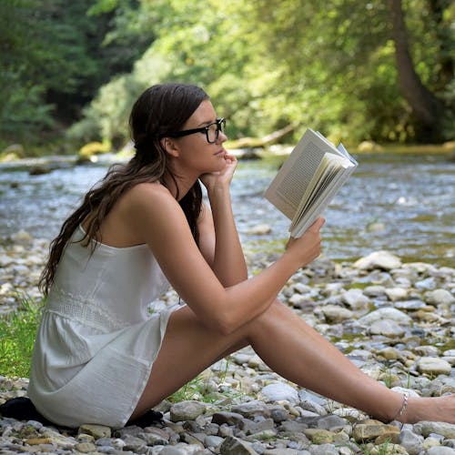 Young woman wearing glasses sitting on stones in front of a lake reading a book. 