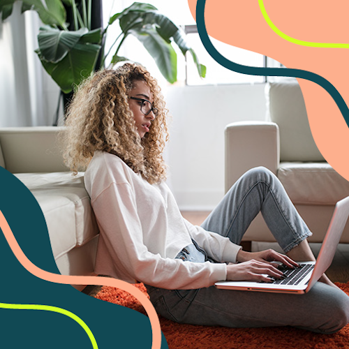 A woman sitting on the living room rug, leaning against the sofa while using her laptop which is balanced on one leg