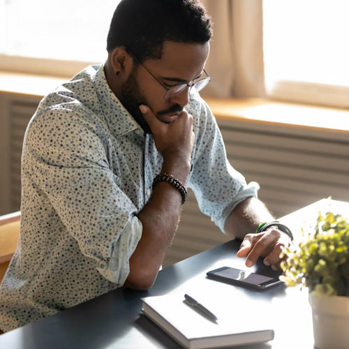 A man with glasses and a collared shirt looking worried stares down at his phone in front of him