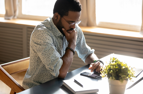 A man with glasses and a collared shirt looking worried stares down at his phone in front of him