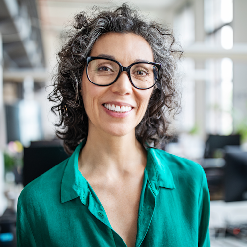 A woman in a green silk button up shirt with black glasses and grey curly hair smiles at the camera. She looks to be in an office setting.