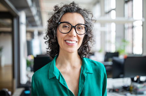 A woman in a green silk button up shirt with black glasses and grey curly hair smiles at the camera. She looks to be in an office setting.