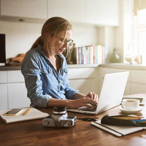 A woman standing at a kitchen table typing on her laptop