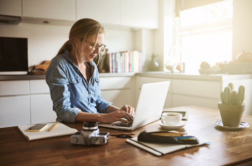 A woman standing at a kitchen table typing on her laptop