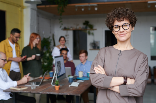 Person with light skin, glasses, and short curly hair stands smiling in the foreground with their arms crossed. A team of professionals works together in the background.