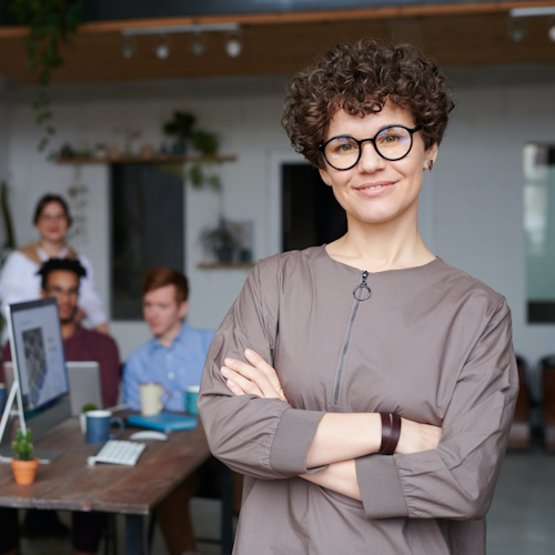 Person with light skin, glasses, and short curly hair stands smiling in the foreground with their arms crossed. A team of professionals works together in the background.