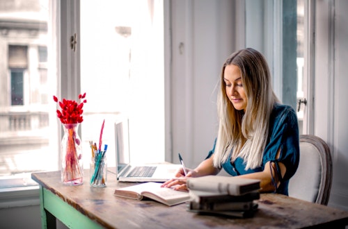 Woman sitting at a laptop writing in a notepad with flowers on her desk. 