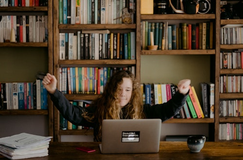 Woman celebrating in front of her laptop with a library in the background, indicating lifelong learning