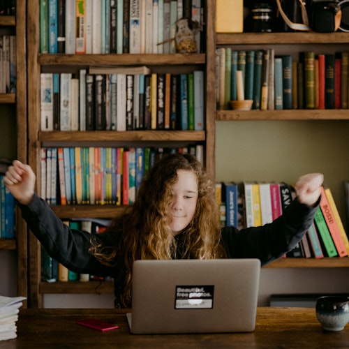 Woman celebrating in front of her laptop with a library in the background, indicating lifelong learning