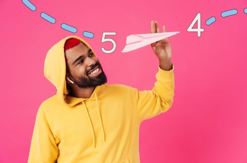 Man in a yellow hoodie holding a paper airplane with a pink background