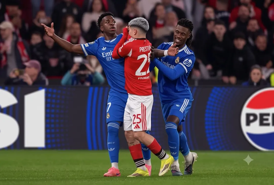 SL Benfica player Gianluca Prestianni (wearing a red jersey, number 25) covers his mouth while speaking closely to Real Madrid player Vinícius Júnior (wearing a blue jersey, number 7), who reacts with his arms outstretched in disbelief, while Eduardo Camavinga attempts to intervene on a football pitch.