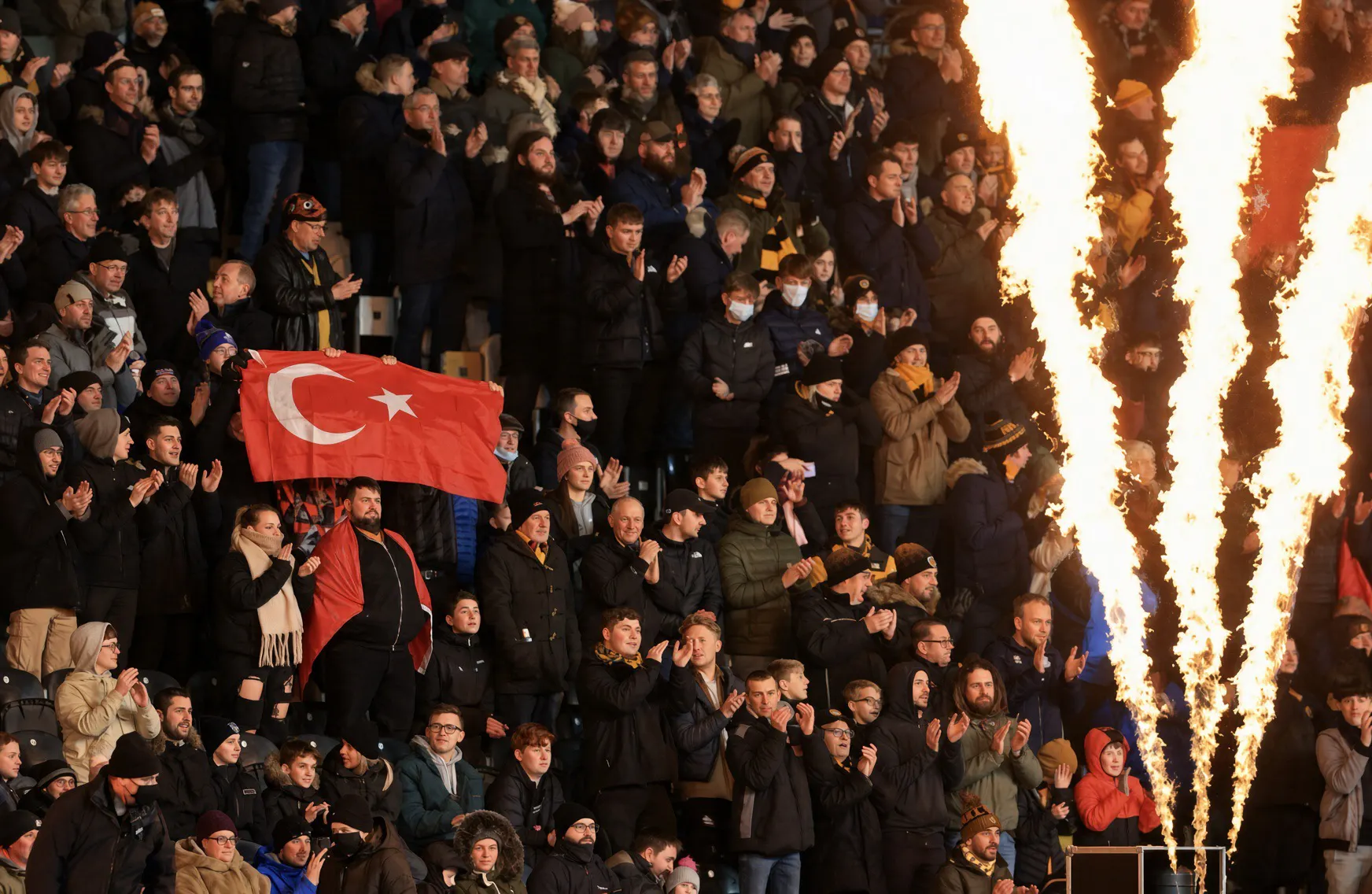 A packed stadium crowd in winter coats claps as a fan holds a large Turkish flag aloft, with tall pyrotechnic flames burning on the right ahead of Hull City vs Everton (FA Cup, MKM Stadium, 8 Jan 2022).