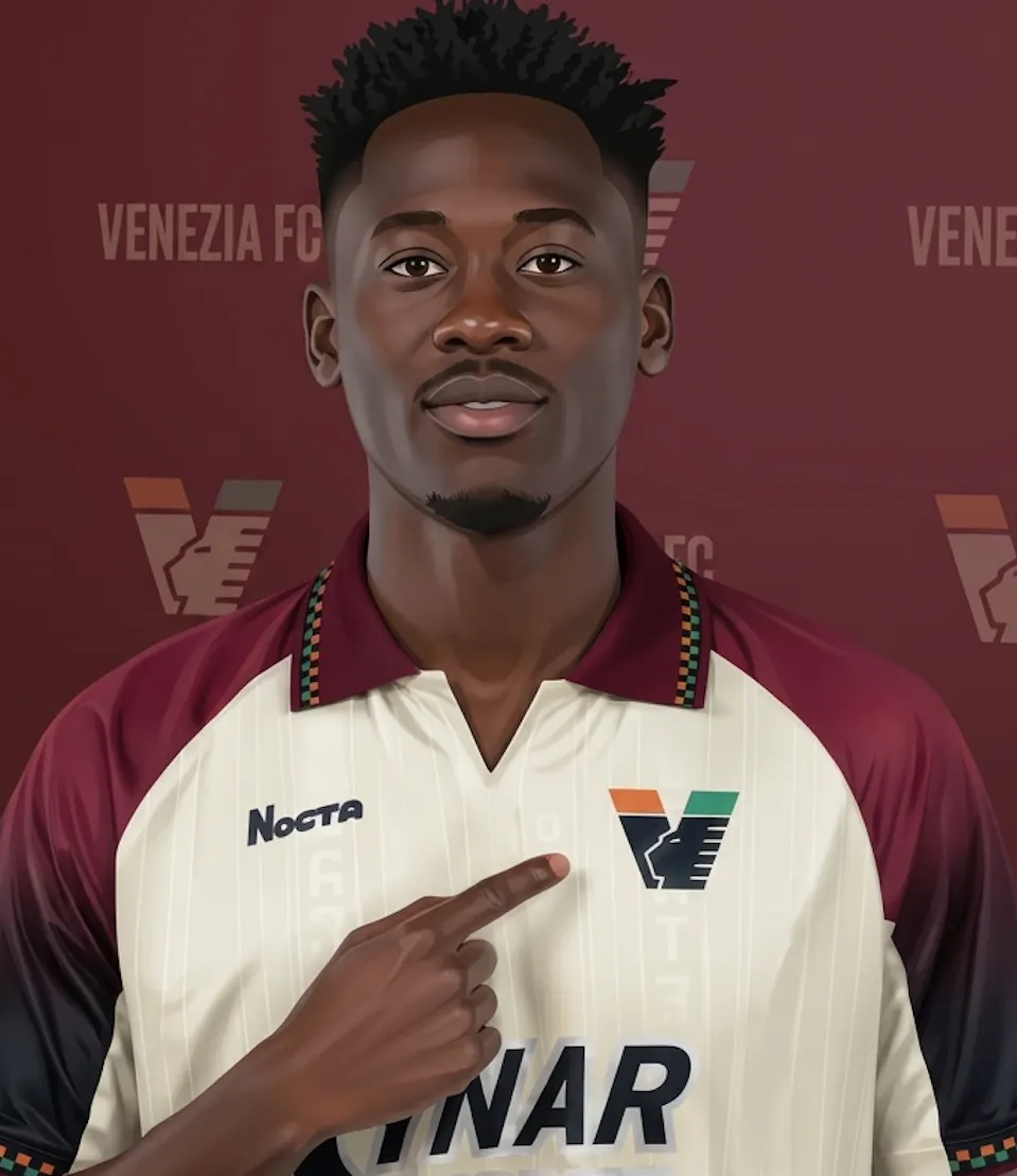 Akor Adams posing during his unveiling for Venezia FC. He wears the club's cream, maroon, and black kit while pointing directly to the Venezia FC club crest on his chest. In the background is a maroon media wall featuring repeating "VENEZIA FC" logos.