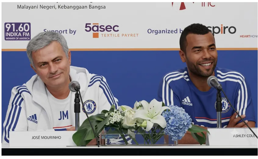 Football manager José Mourinho and former player Ashley Cole sitting side-by-side at a press conference table, both smiling while wearing Chelsea FC athletic gear with microphones in front of them.