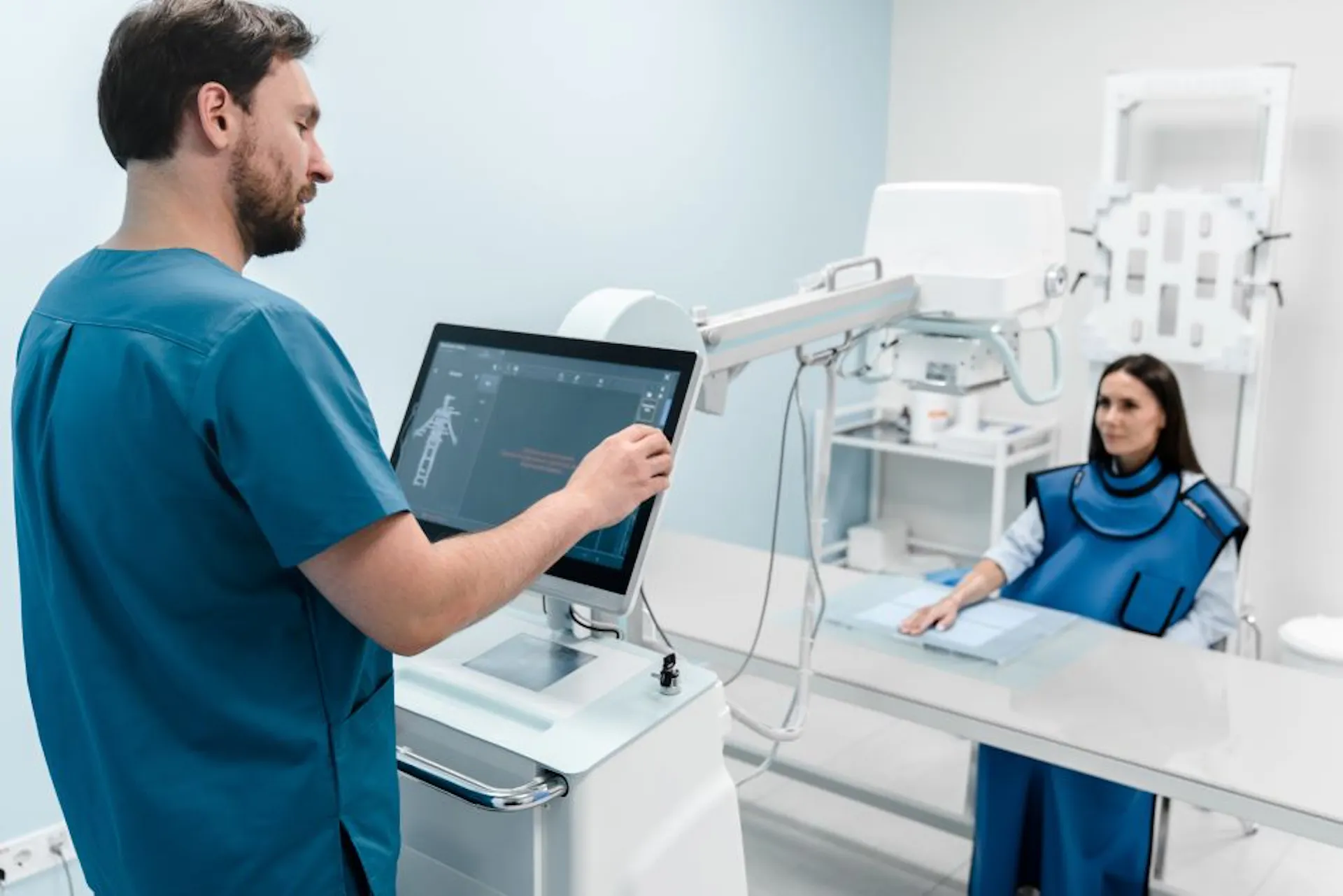 Male radiologist doing X-ray of female patient's hand on X-ray machine at. Patient wearing a protective lead apron. 
