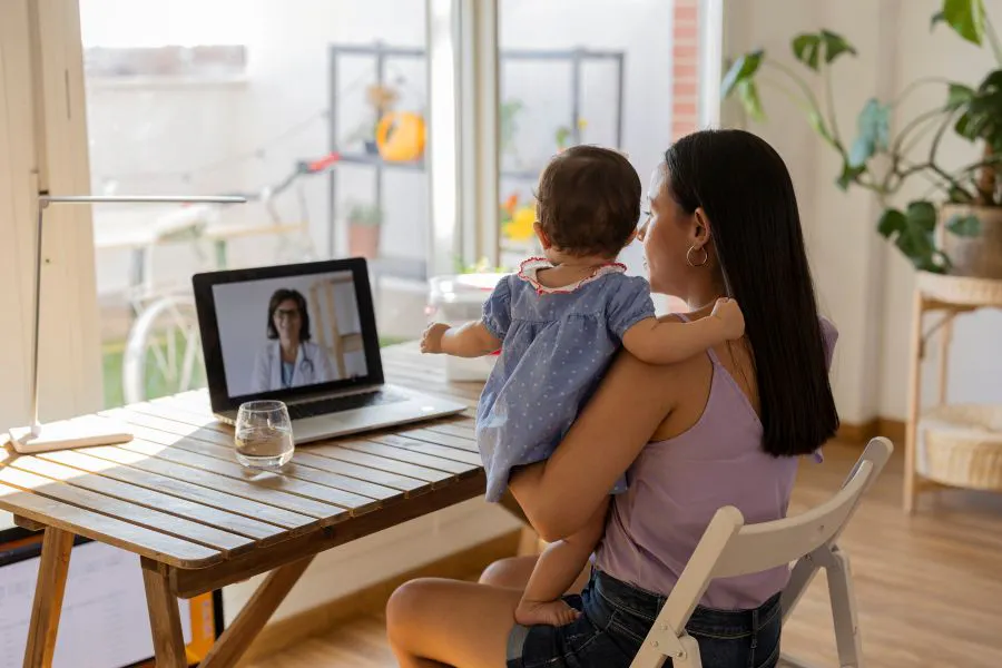 A mother with her daughter in her arms sitting on a video call with the doctor - virtual appointment