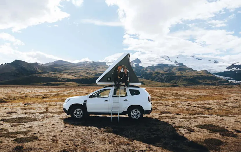 2 travelers sitting in a pop up tent on top of a white Dacia with Icelandic mountains in the background