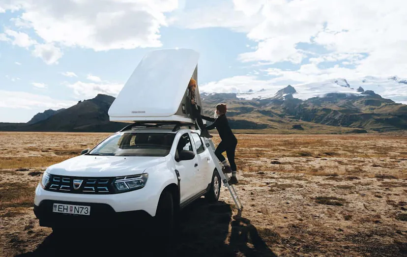 Traveler climbing up to a pop up tent on top of a white Dacia with Icelandic mountains in the background