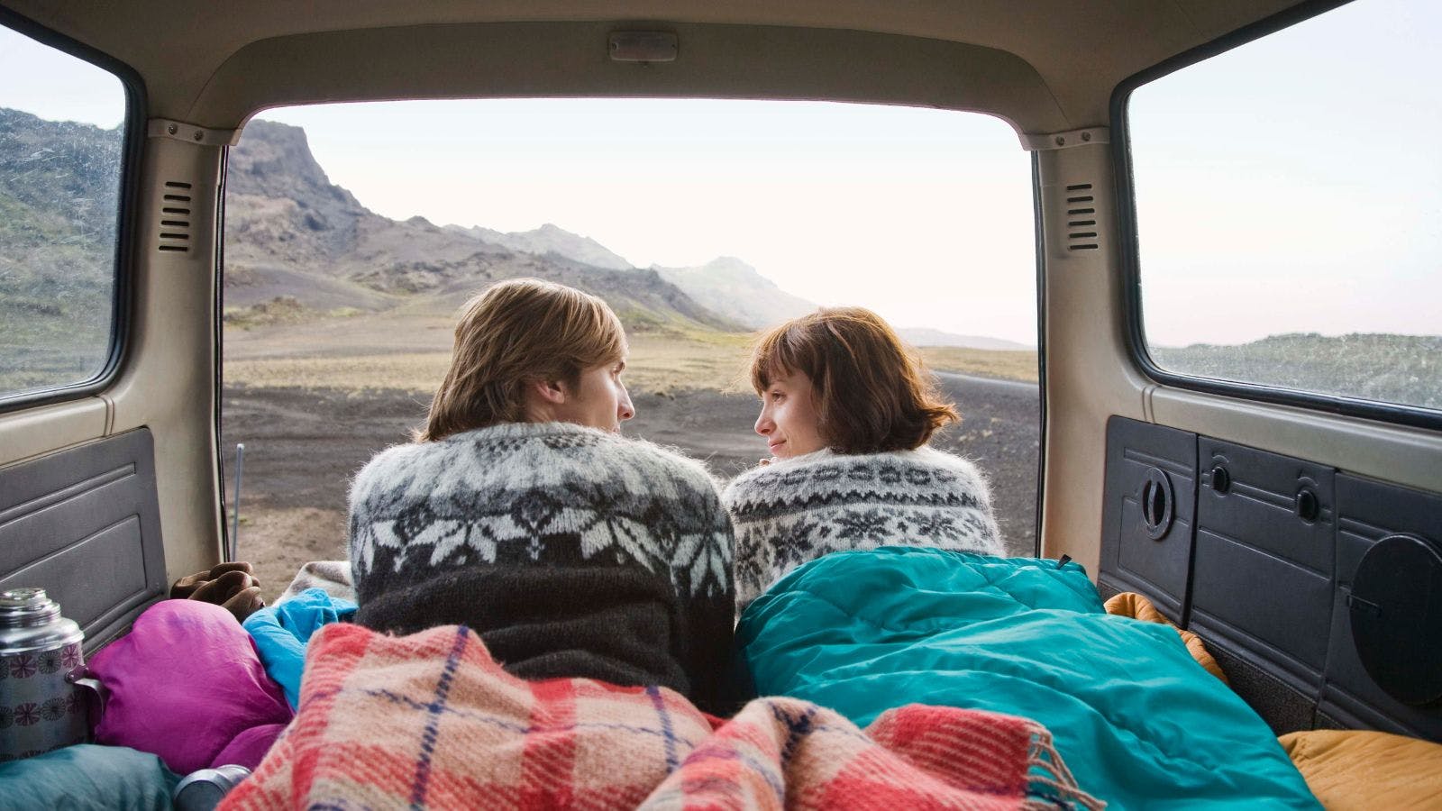 Couple camping in Iceland in the back of a camper van, enjoying the scenery beneath blankets.