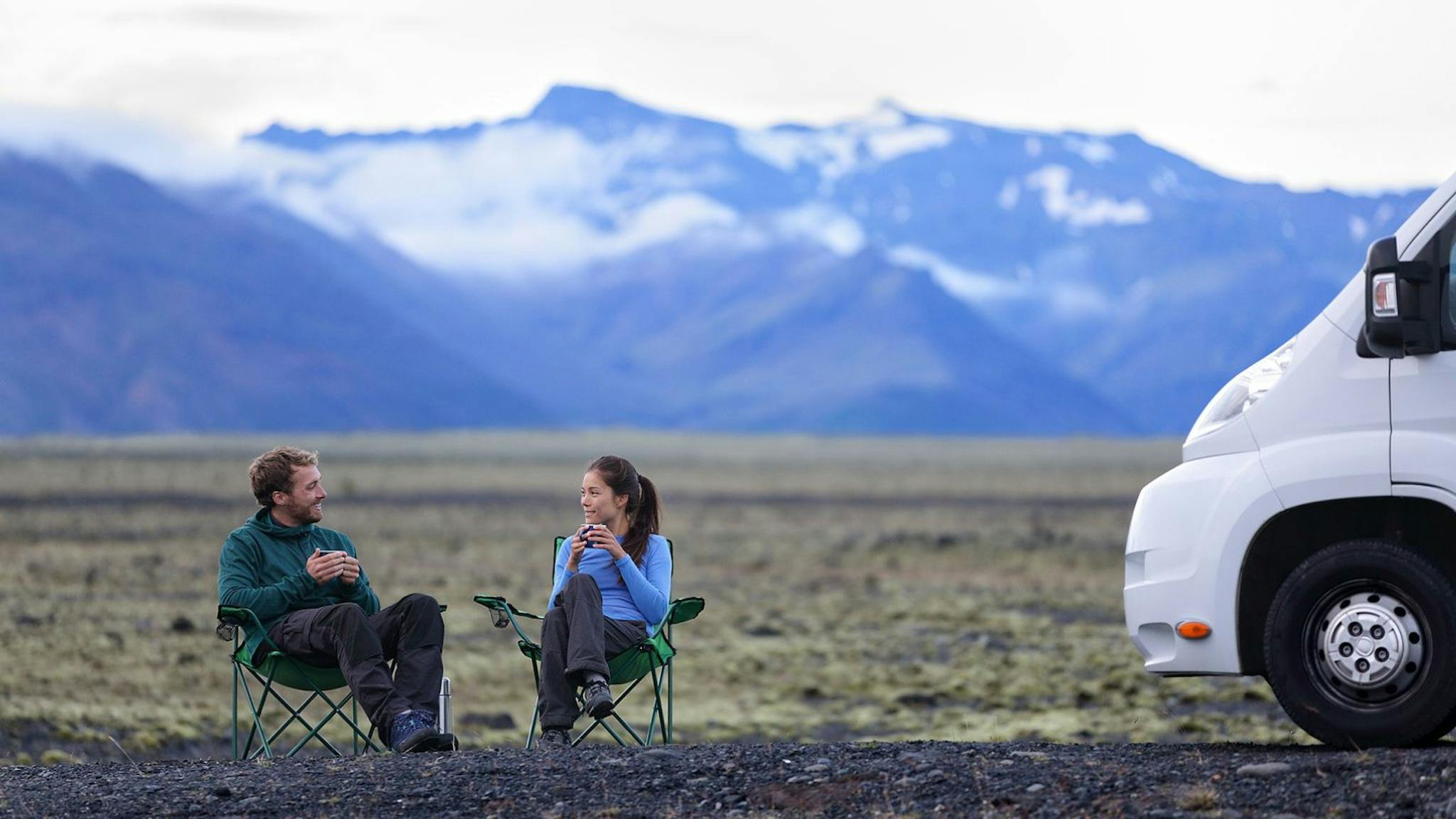 A couple in Iceland sipping coffee outside their campervan with mountains in the background.