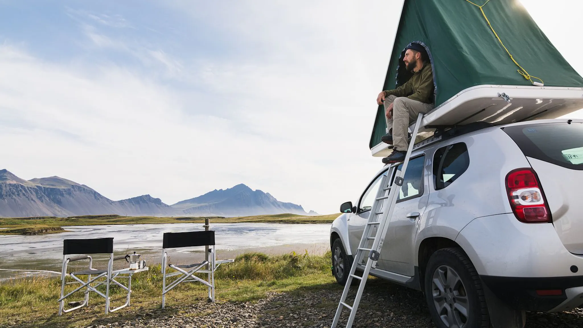 Man sitting in a rooftop tent camper overlooking a river in Iceland.