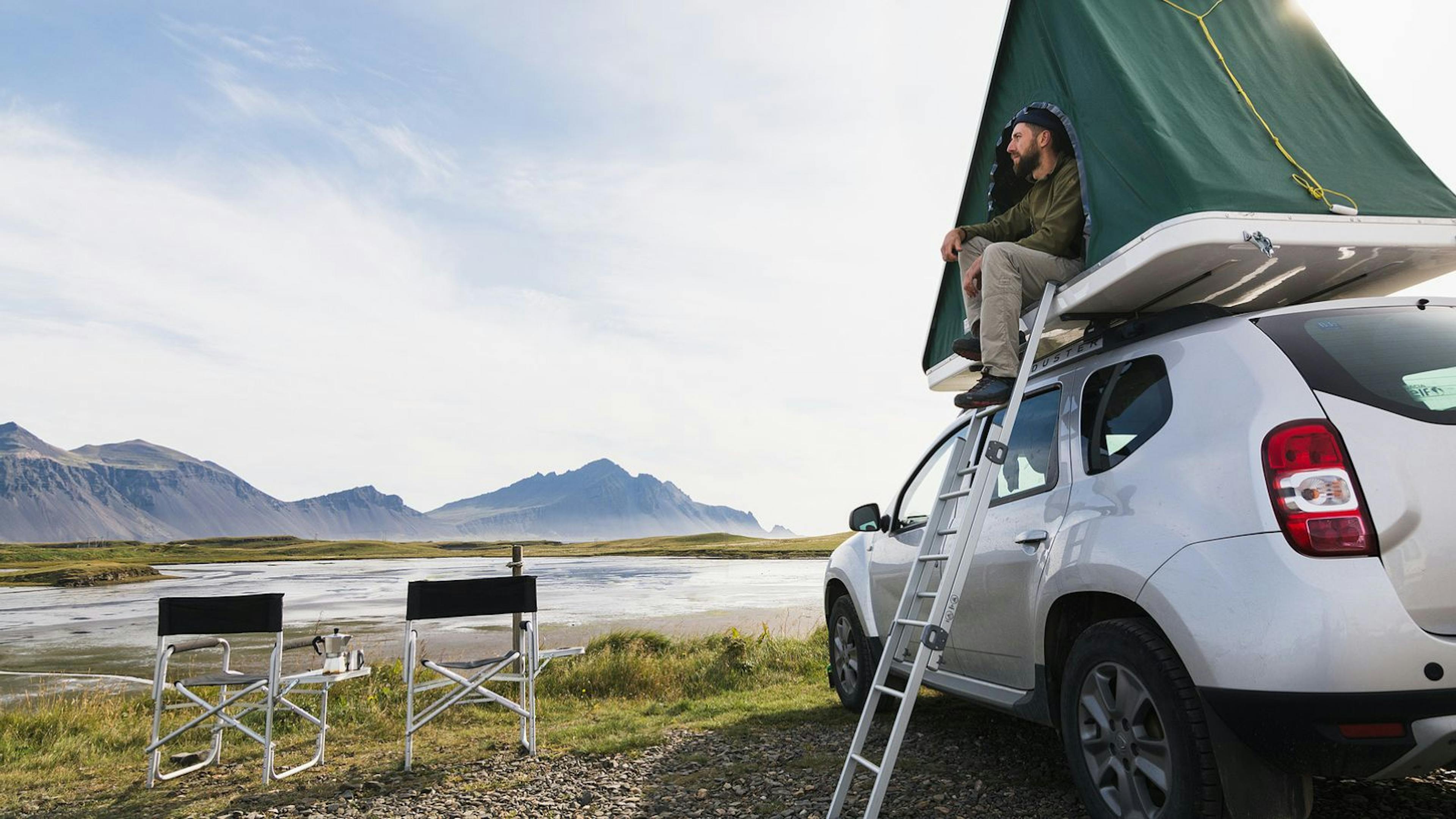 Man sitting in a rooftop tent camper overlooking a river in Iceland.