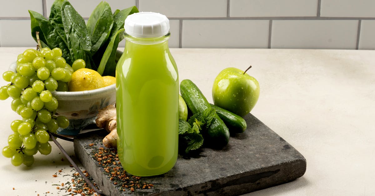 bottle of green juice with ingredients grapes, spinach, green tea, cucumber, mint, lemon and green apple on a countertop