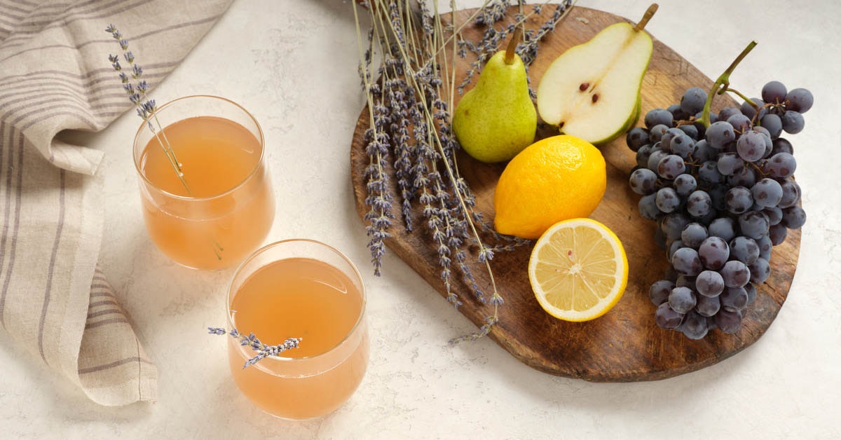two glasses of grape juice on a white table surrounded by recipe ingredients lemon, pear, grapes and lavender