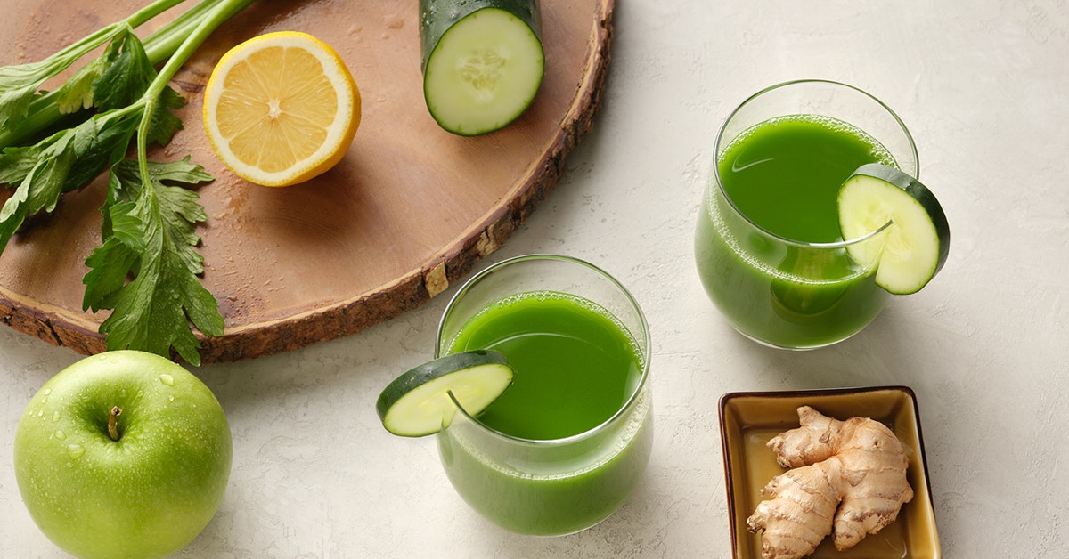 two glasses of green juice surrounded by the ingredients cucumber, lemon, green apple and kale