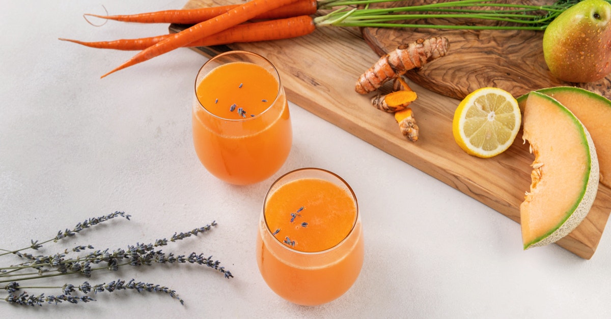 two glasses of cantaloupe melon juice with carrots, turmeric, lemon, pear and lavender on a wooden cutting board