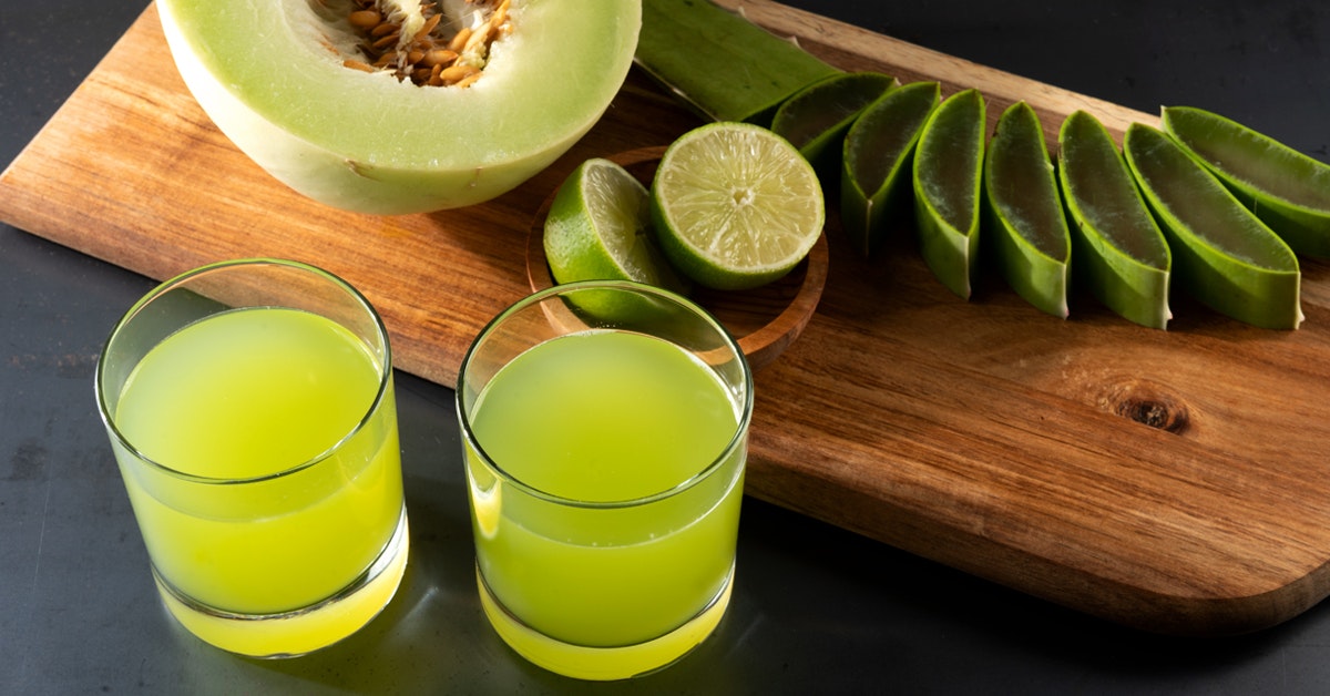 two glasses of renew and repair aloe juice next to a wooden cutting board of ingredients honeydew, aloe vera and lime