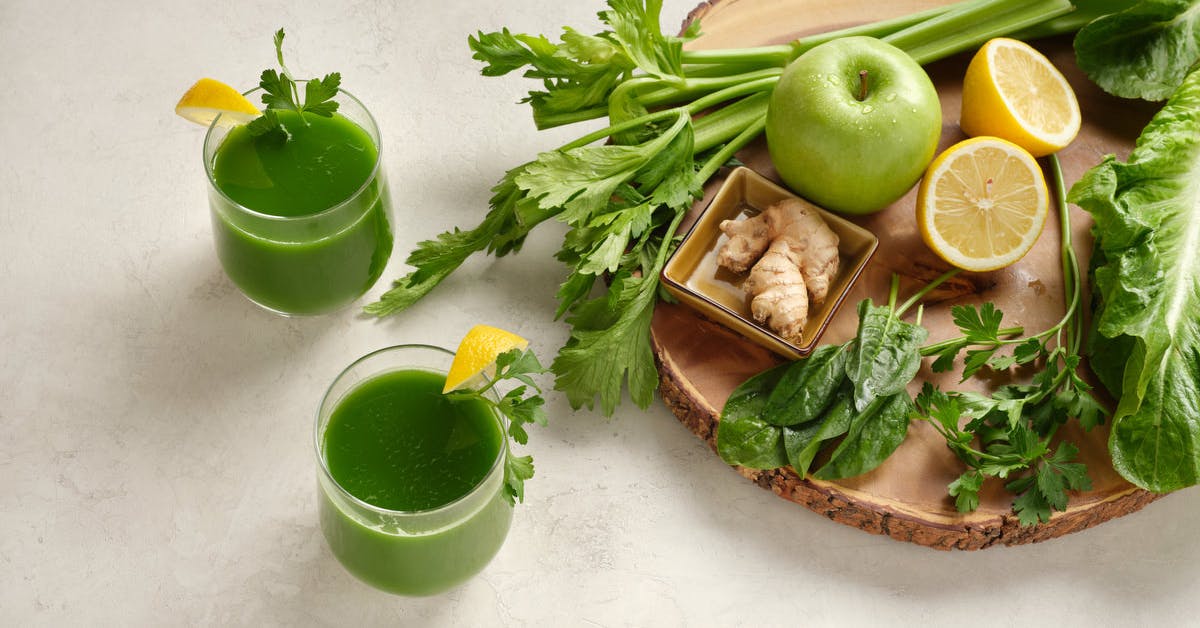 two glasses of green juice with the ingredients, spinach, celery, green apple, ginger, lemon and parsley on a wood cutting board