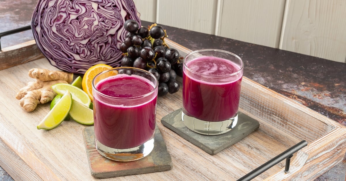 red cabbage juice with grape on a wooden serving table surrounded by recipe ingredients cabbage, grape, ginger, lime and orange