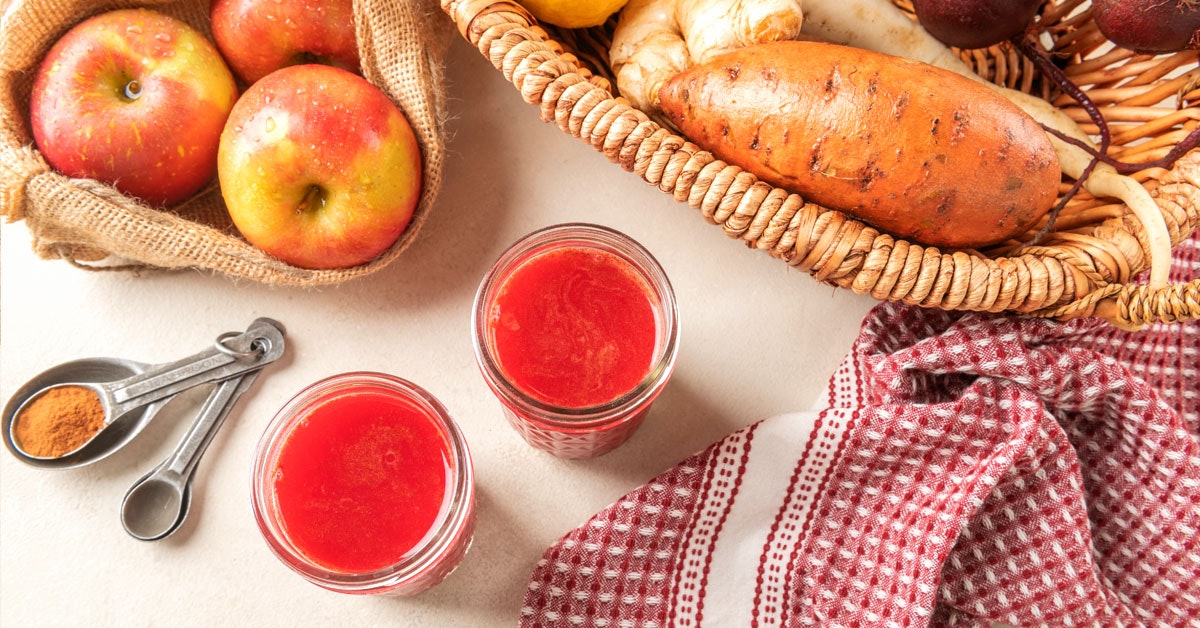 two glasses of cold pressed fall harvest juice on a white table next to a basket of ingredients
