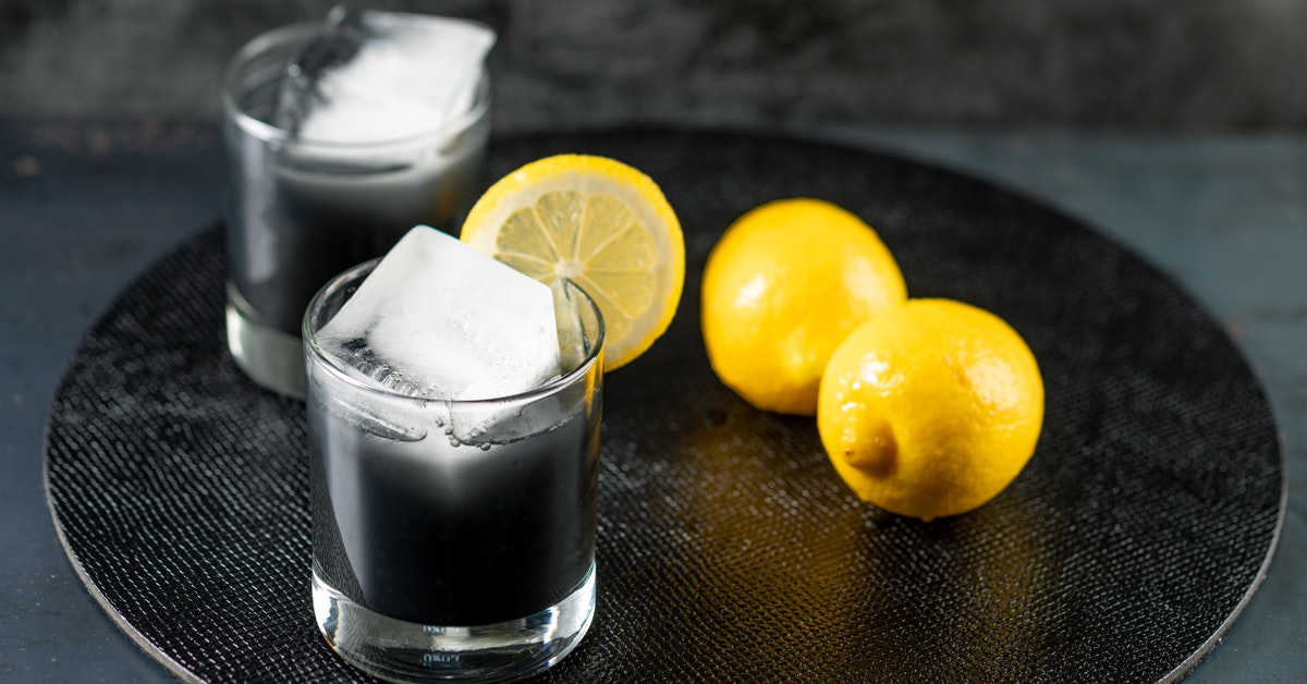 two glasses of lemonade with activated charcoal sitting on a black place mat next to lemons