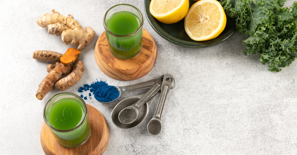 two servings of green juice shots on a table with ginger, turmeric, lemon, kale and spirulina 