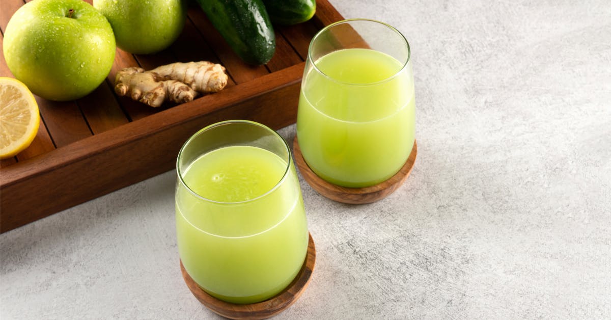 two glasses of glowing skin green juice on wooden coasters next to a tray of ingredients