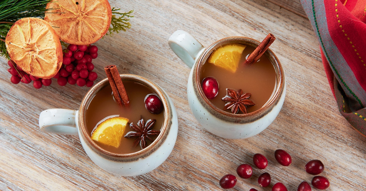 two mugs of spiked warm apple cider cocktail with cinnamon sticks, orange, cranberry, and clove