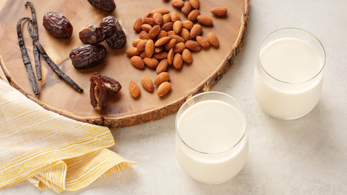 two glasses of cold pressed almond milk surrounded by dates, vanilla and almonds on a wood cutting board.