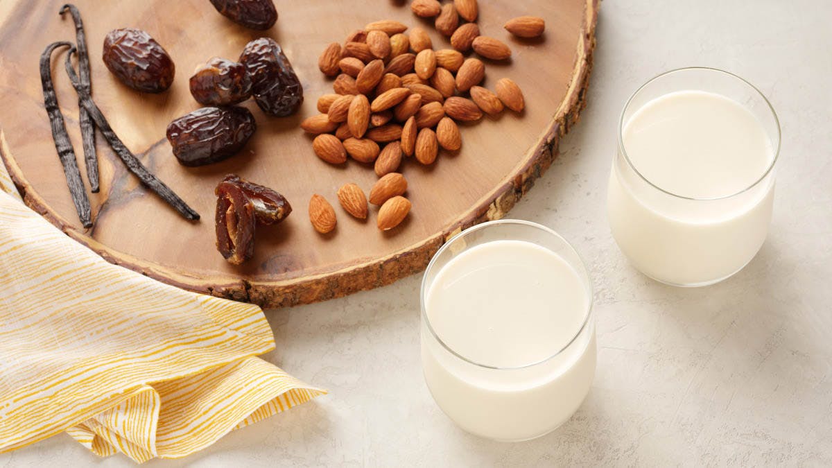 two glasses of cold pressed almond milk surrounded by dates, vanilla and almonds on a wood cutting board