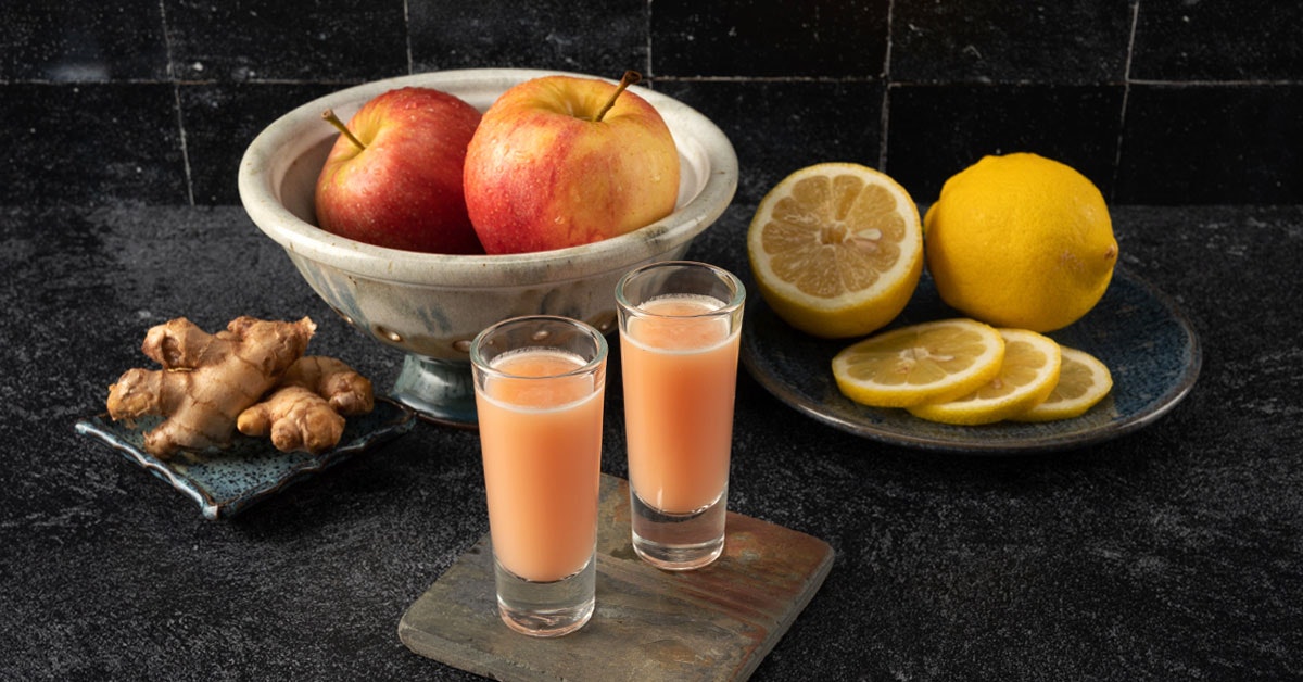 two healthy ginger cold pressed juice shots next to lemon, apple and ginger on a black countertop