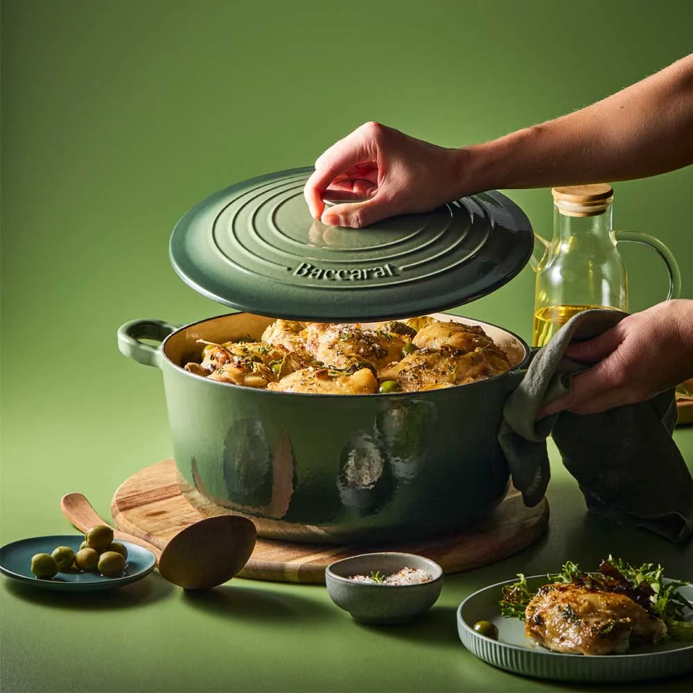 A pair of hands is lifting the lid off a sage Dutch oven filled with cooked chicken and herbs. The pot is resting on a wooden board against a solid sage green background.