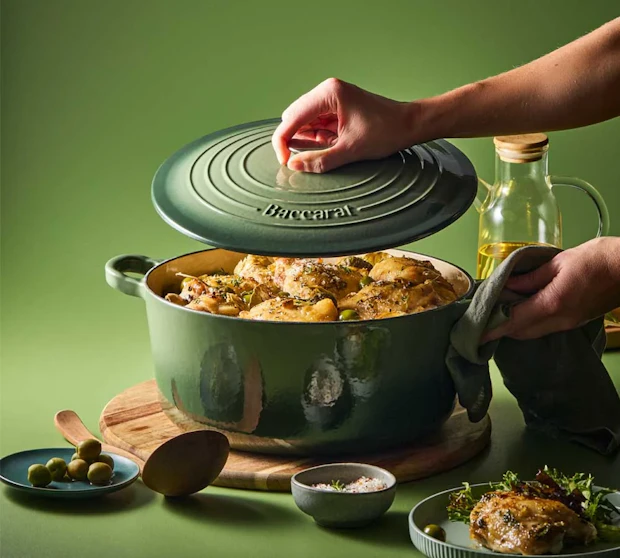 A pair of hands is lifting the lid off a sage Dutch oven filled with cooked chicken and herbs. The pot is resting on a wooden board against a solid sage green background.