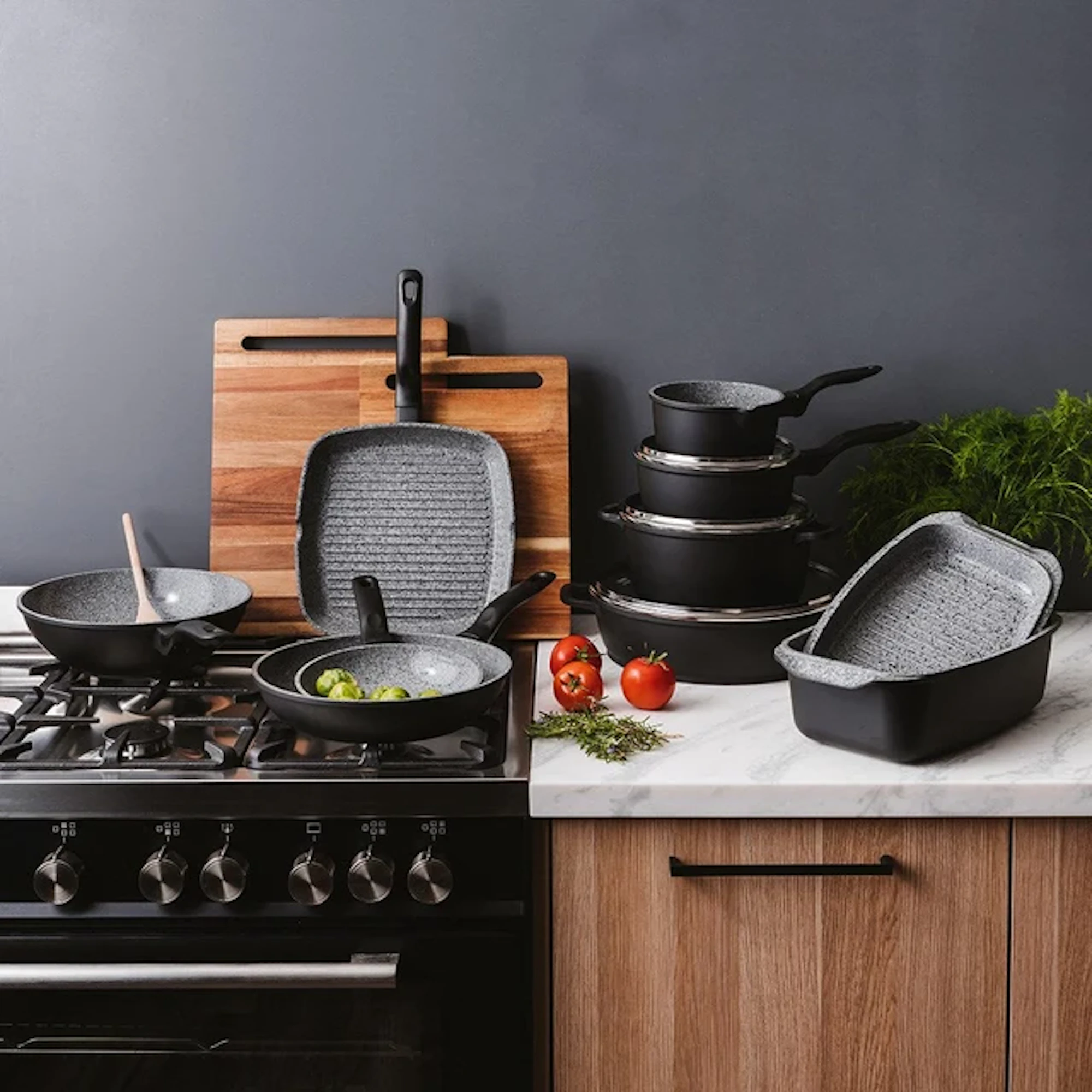 A black cookware set is displayed in a kitchen with pots on a stove, pans, cutting boards, and tomatoes on the counter.