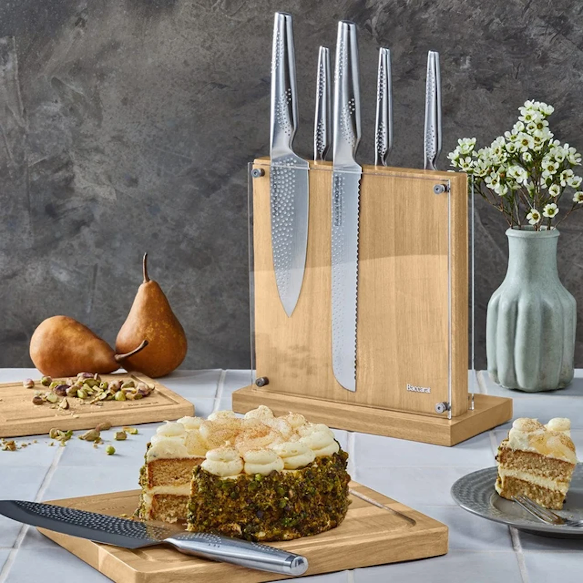 A frosted cake is on a wooden cutting board with a knife beside it, while a knife block, pears, and a vase of flowers sit in the background.
