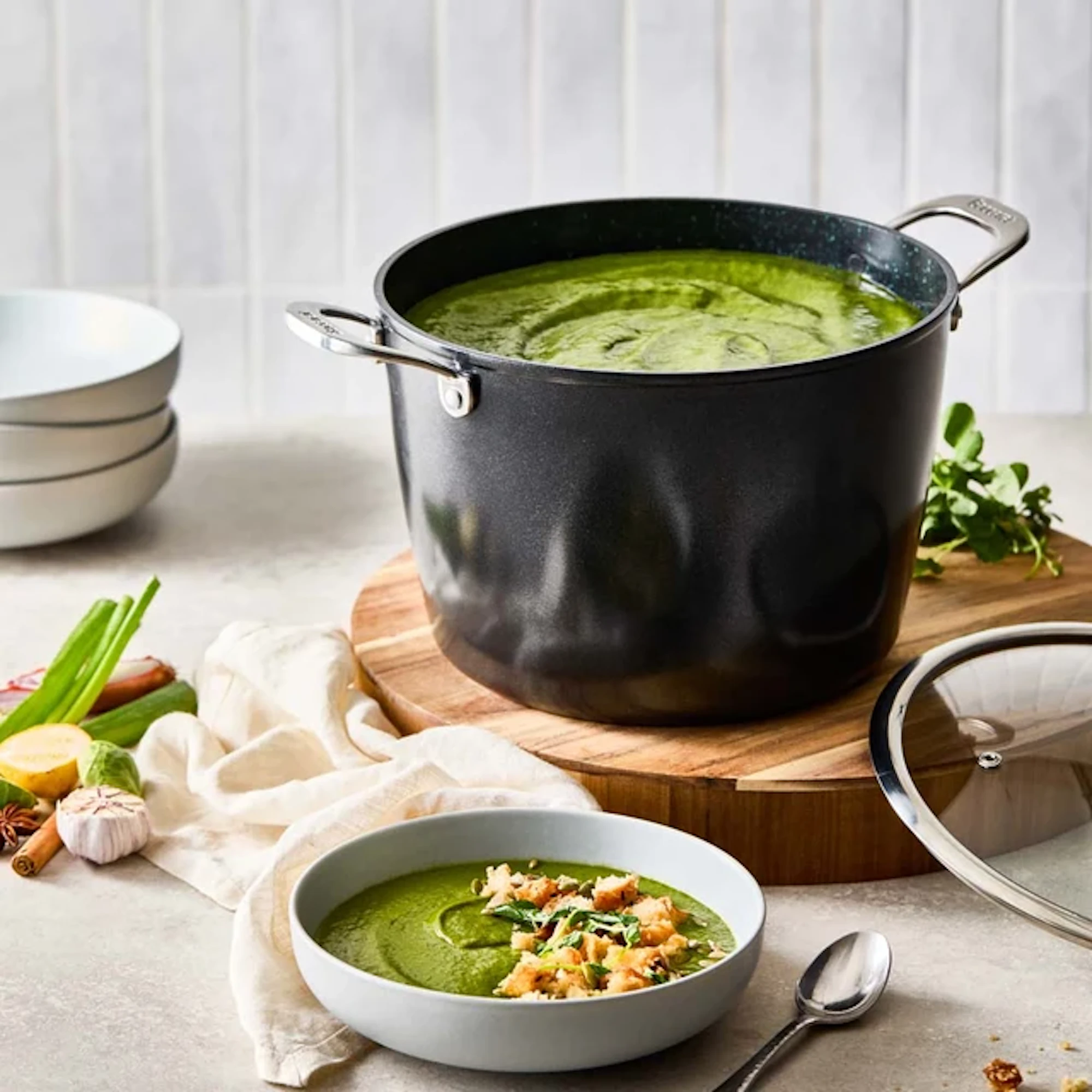 A large black pot is resting on a wooden board and is filled with green soup. In the foreground, a light-colored bowl contains a serving of the green soup garnished with croutons and herbs next to a spoon.