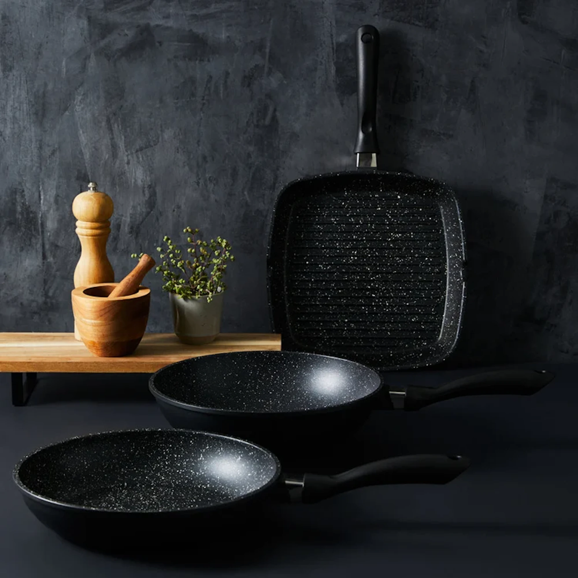 Three black non-stick pans are displayed on a dark surface against a textured black background, with wooden kitchen accessories and a small potted plant on a wooden shelf.