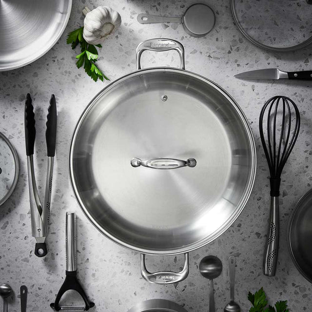 A stainless steel pan is centered on a speckled surface, surrounded by kitchen utensils and cookware.