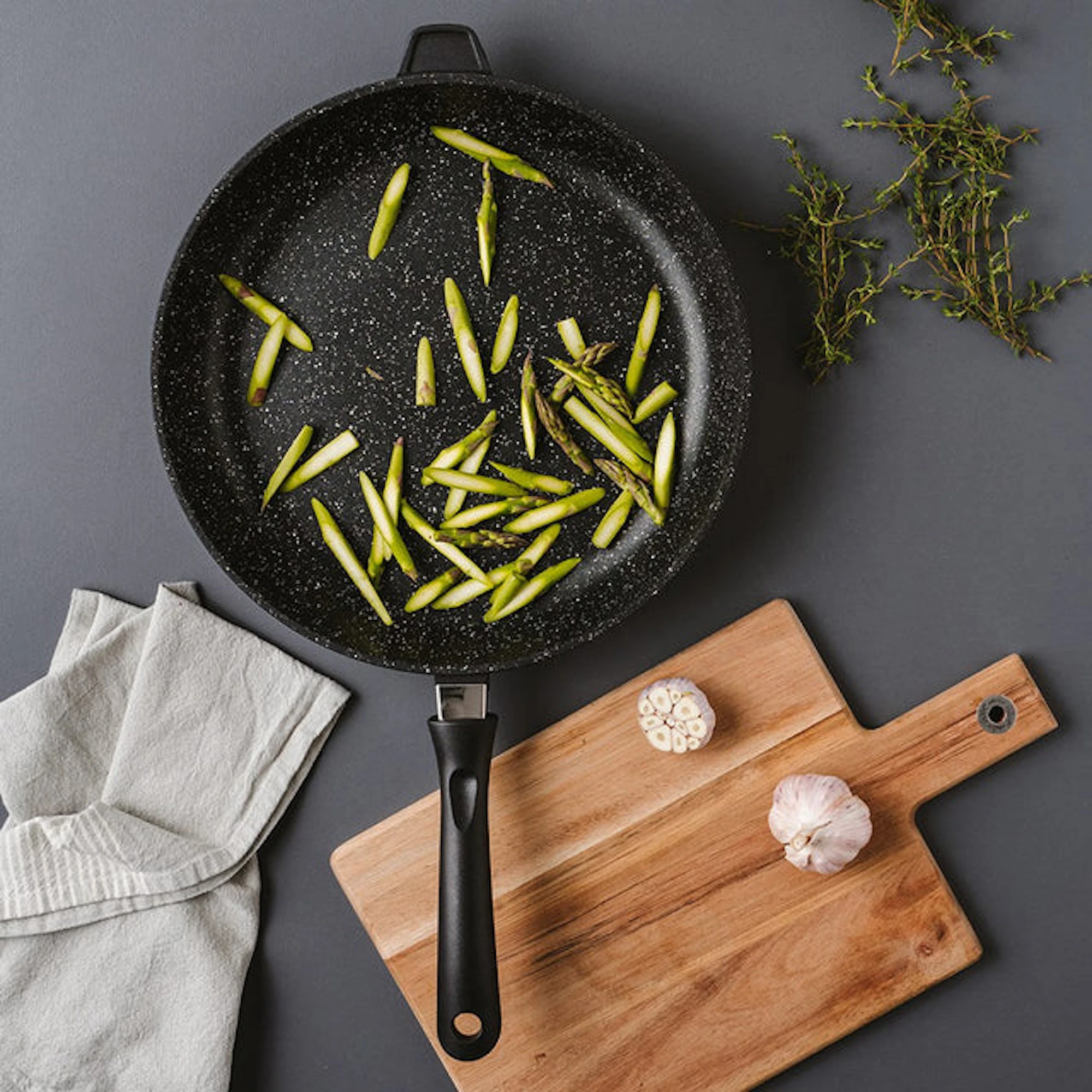 A black granite frying pan is positioned on a dark gray surface, containing scattered pieces of chopped asparagus