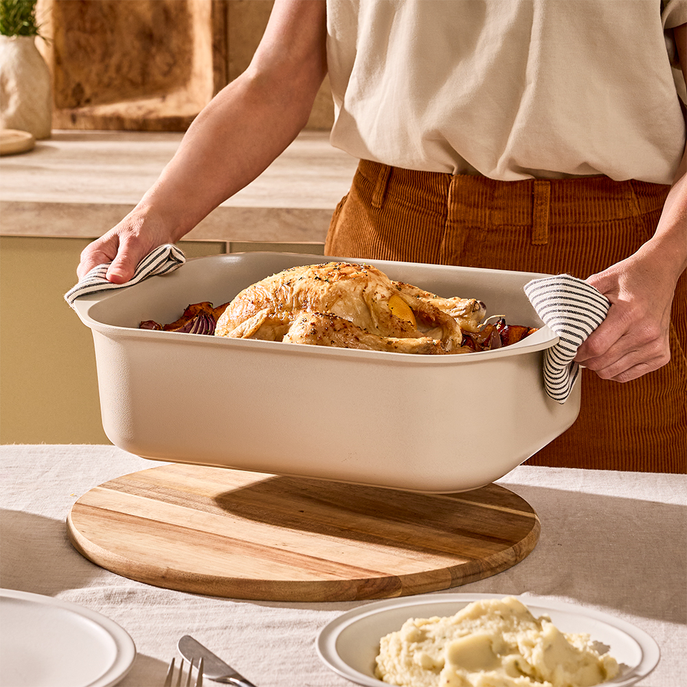 Person carrying the NOOK Cream roaster with a roast chicken in it, placing it on a kitchen table